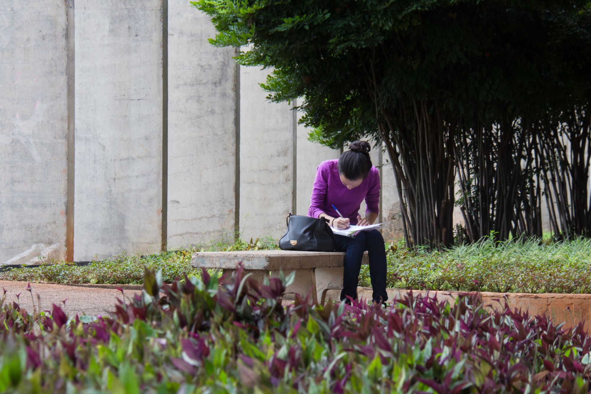 Estudante sentada em um banco de concreto no jardim do campus, escrevendo em um caderno apoiado no colo